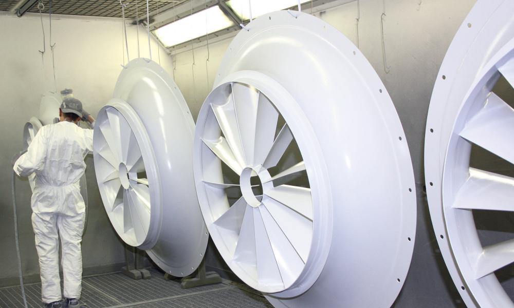 Person in weißem Schutzanzug betrachtet große weiße Ventilatorflügel in einer Fabrikumgebung.